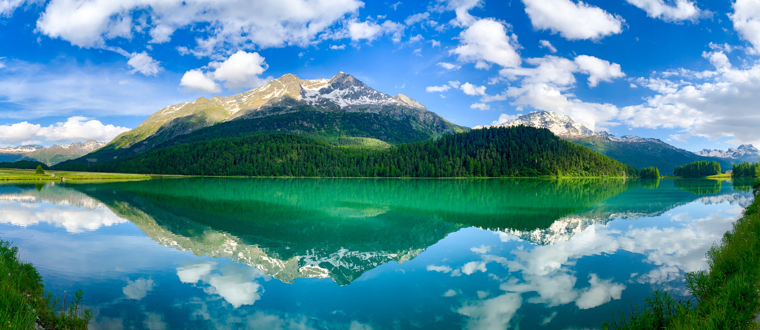Sitting by Lake Silvaplana with a view of the turquoise-blue mountain lake water with lightly snow-capped mountains and green fir forest in the background, reflected in the mountain lake. The sky is blue and dotted with a few white clouds. A picture full of natural beauty.